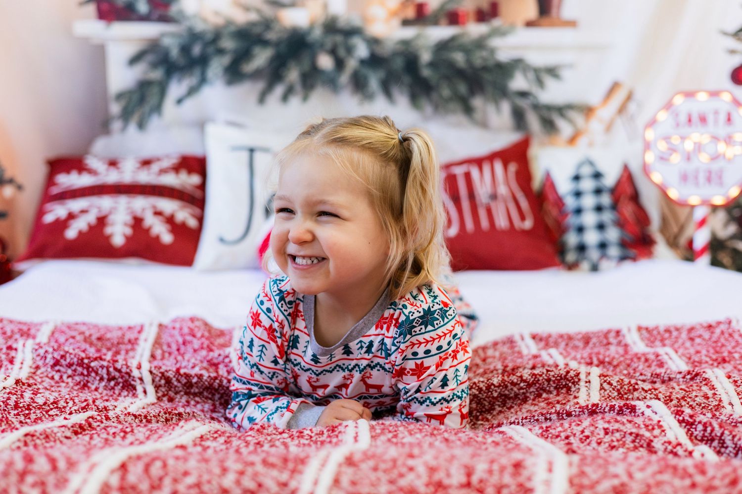 Adorable blonde toddler girl smiling on a red Christmas-themed bed with festive holiday decorations.