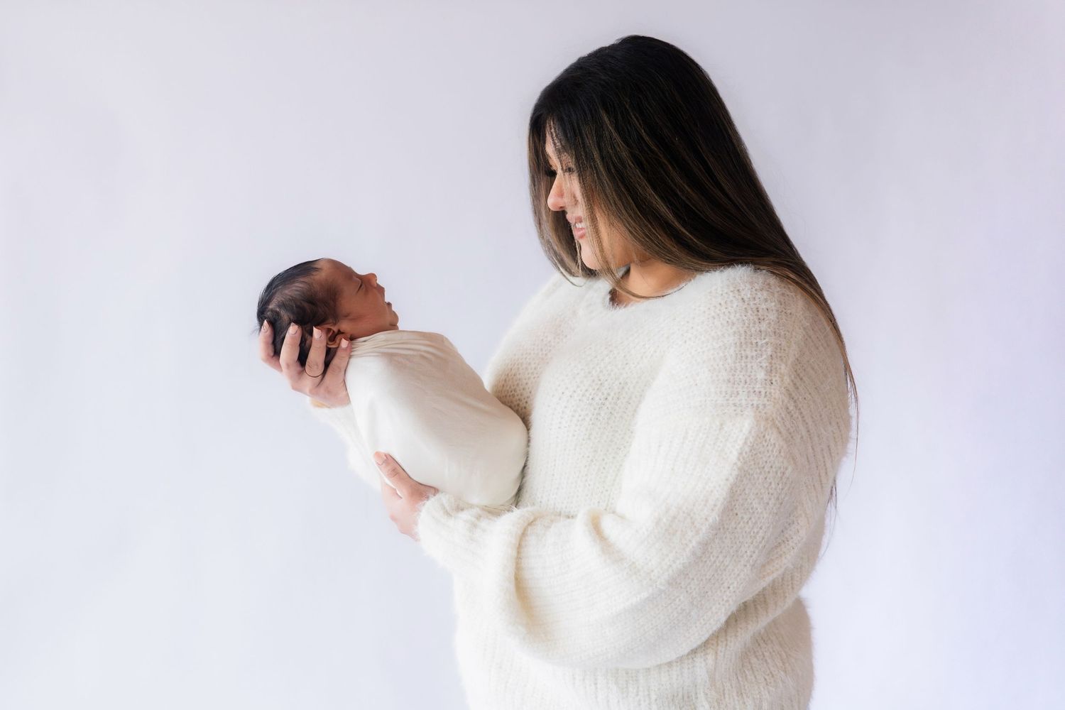 A mother in a cream sweater tenderly holds her swaddled newborn baby against a white background.