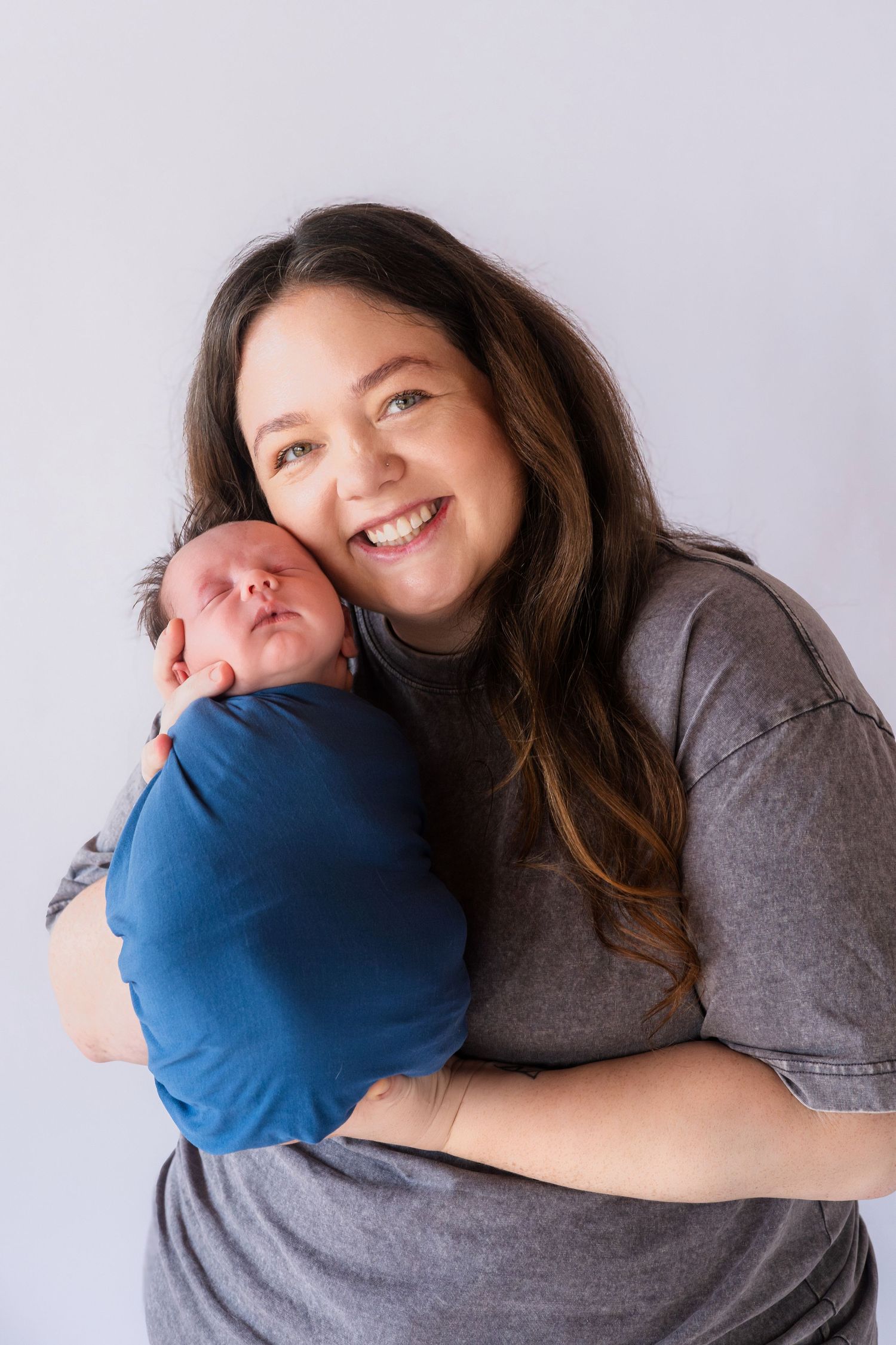 Smiling brunette mother in gray shirt holds her blue-swaddled newborn baby close to her cheek.