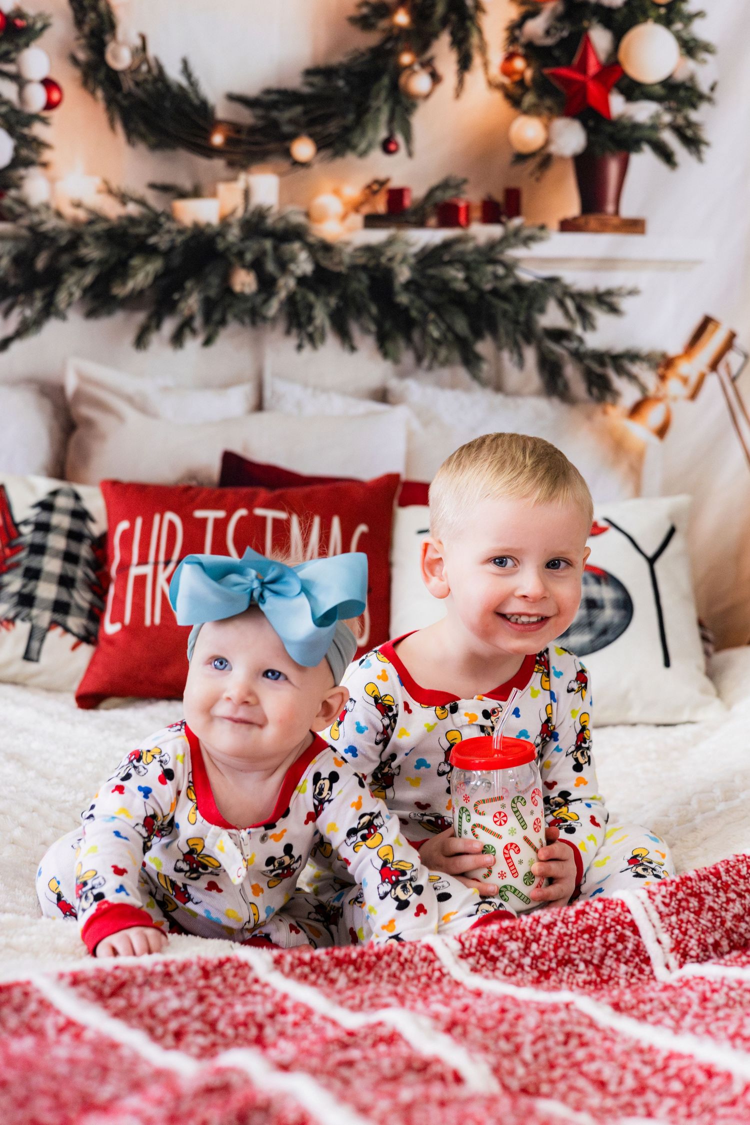 Two young siblings in matching Mickey Mouse pajamas sitting on bed with Christmas decorations behind them.