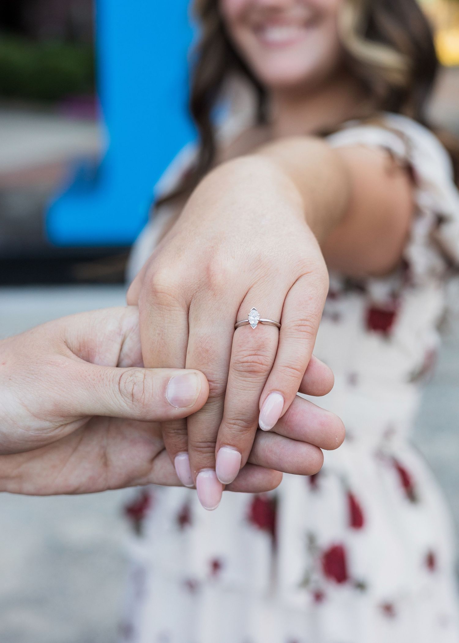 Close-up of womans hand displaying oval diamond engagement ring, held by partner with smiling bride in background.