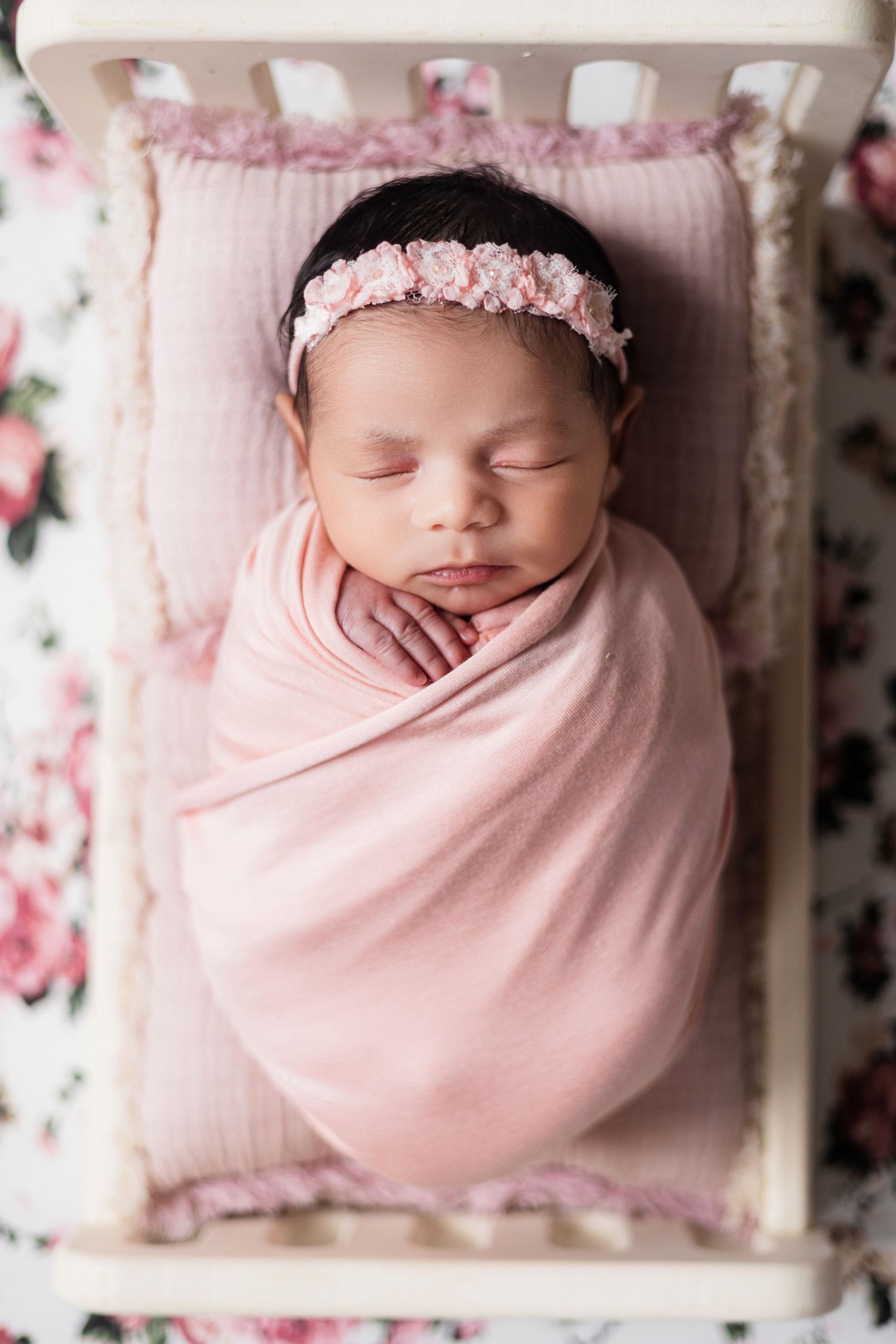 Newborn baby girl swaddled in pink wrap with floral headband sleeping in small white wooden crib on floral mat.