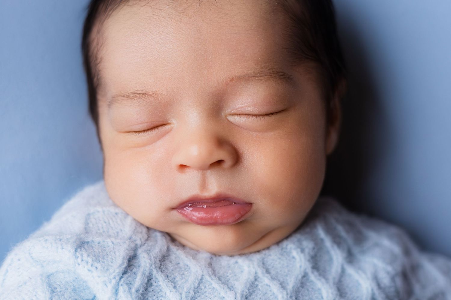 Close-up portrait of sleeping newborn baby with dark hair, wrapped in soft blue knit blanket.