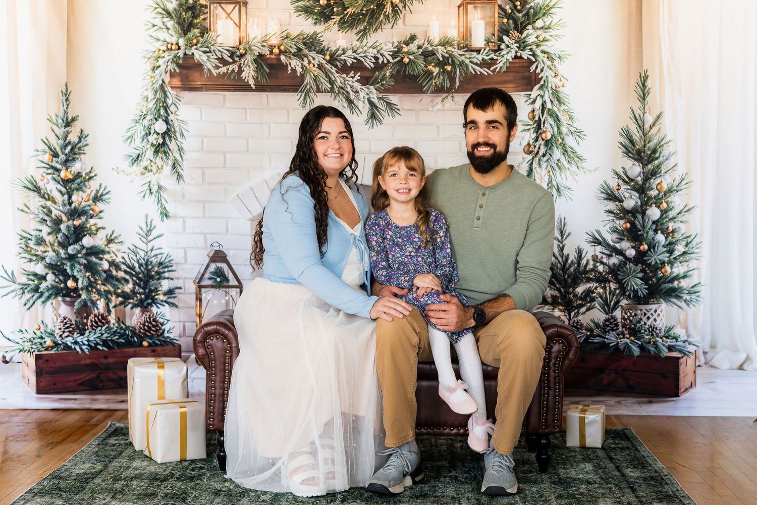 A happy family of three poses in front of a festive Christmas fireplace with decorated trees and gifts.