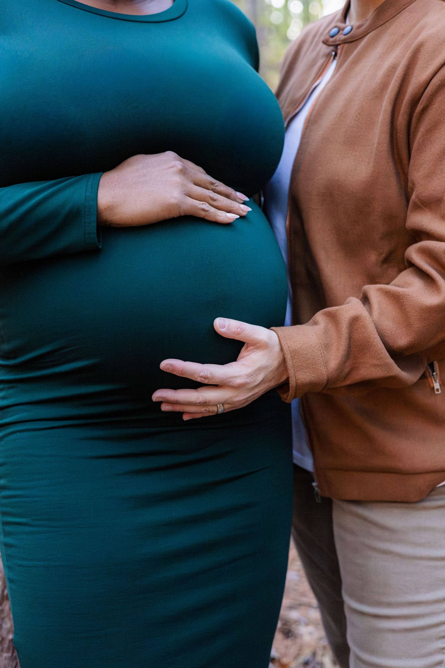 Pregnant woman in teal dress with partner's hand on baby bump outdoors.