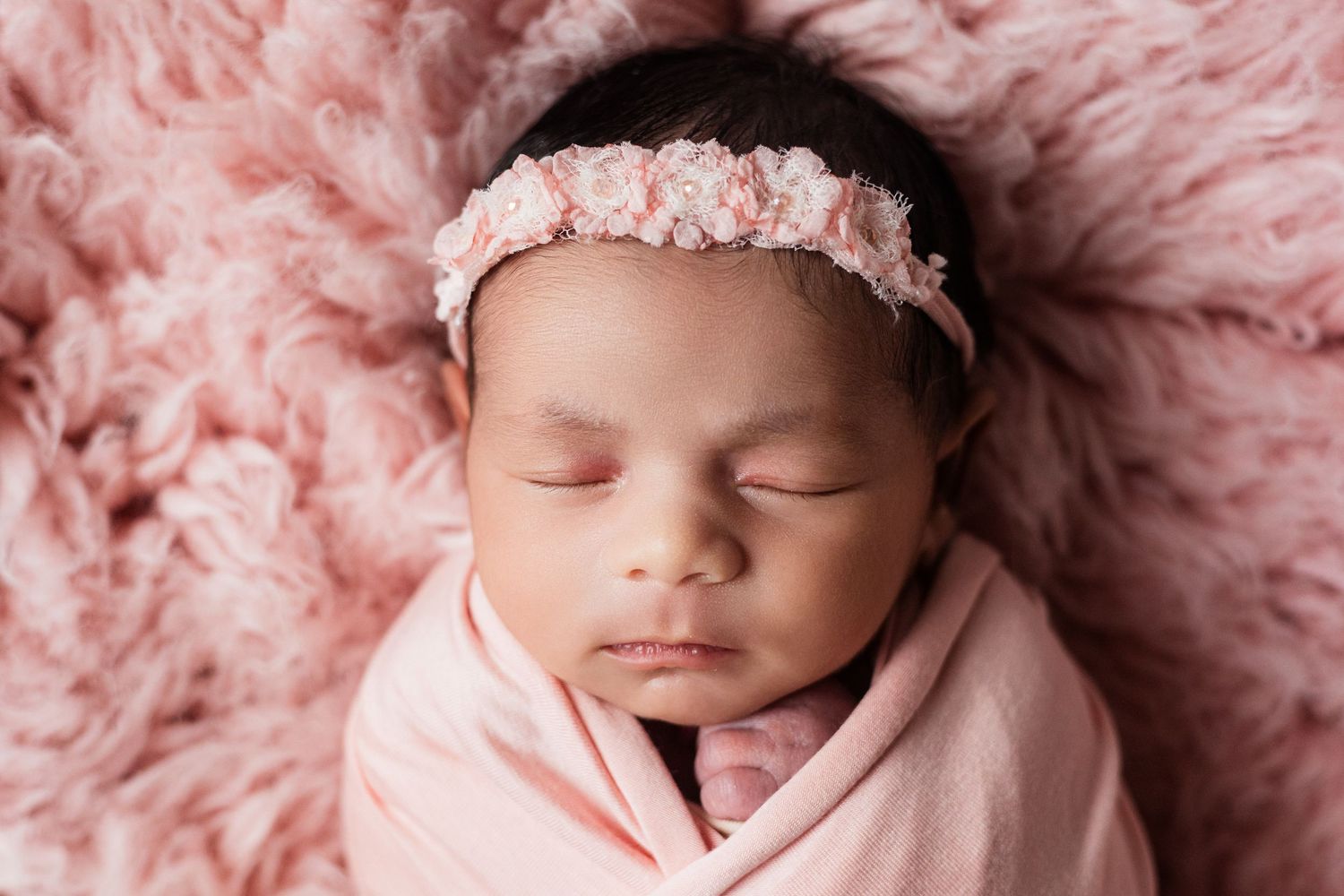 Peaceful newborn baby girl sleeping swaddled in pink wrap with floral headband on soft pink fluffy blanket.