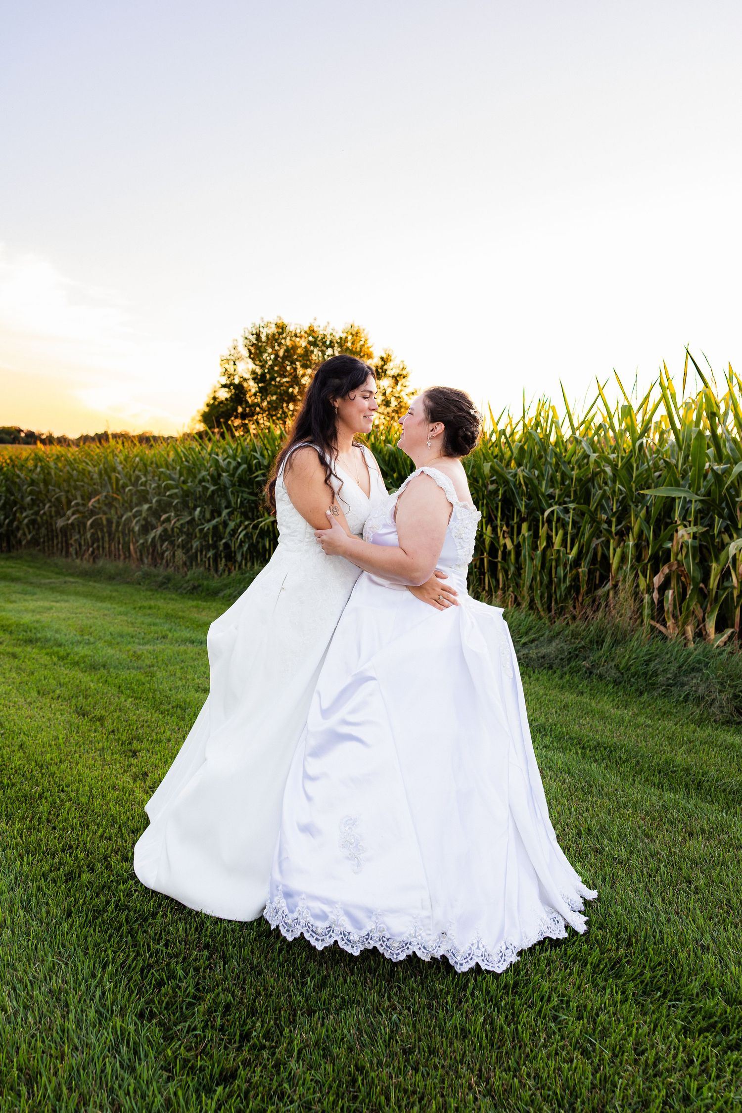 Two brides in white gowns share a romantic moment at sunset beside a lush cornfield on their wedding day.