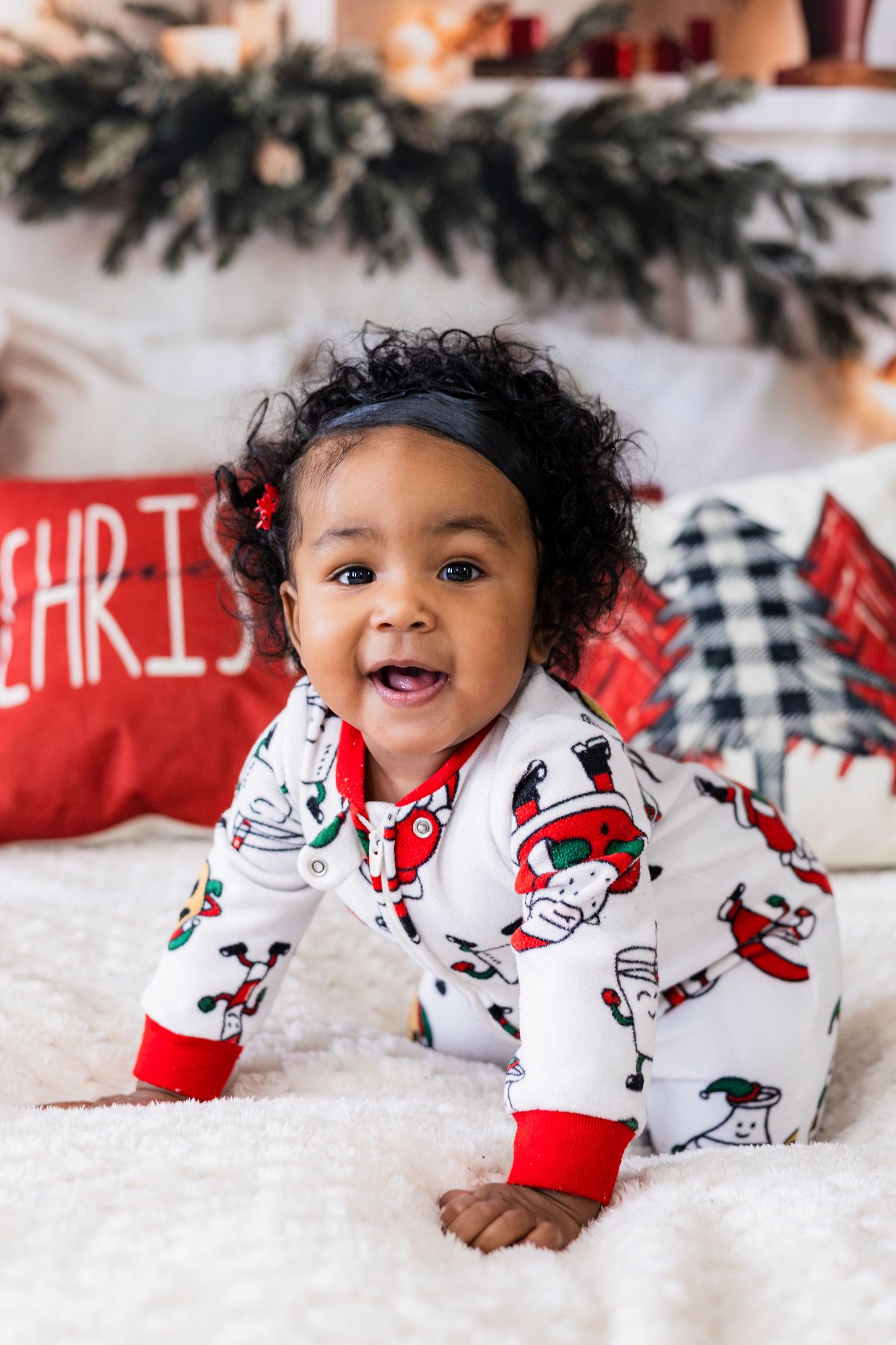 Happy baby girl in Santa Claus pajamas crawling on white blanket with Christmas decorations in background.