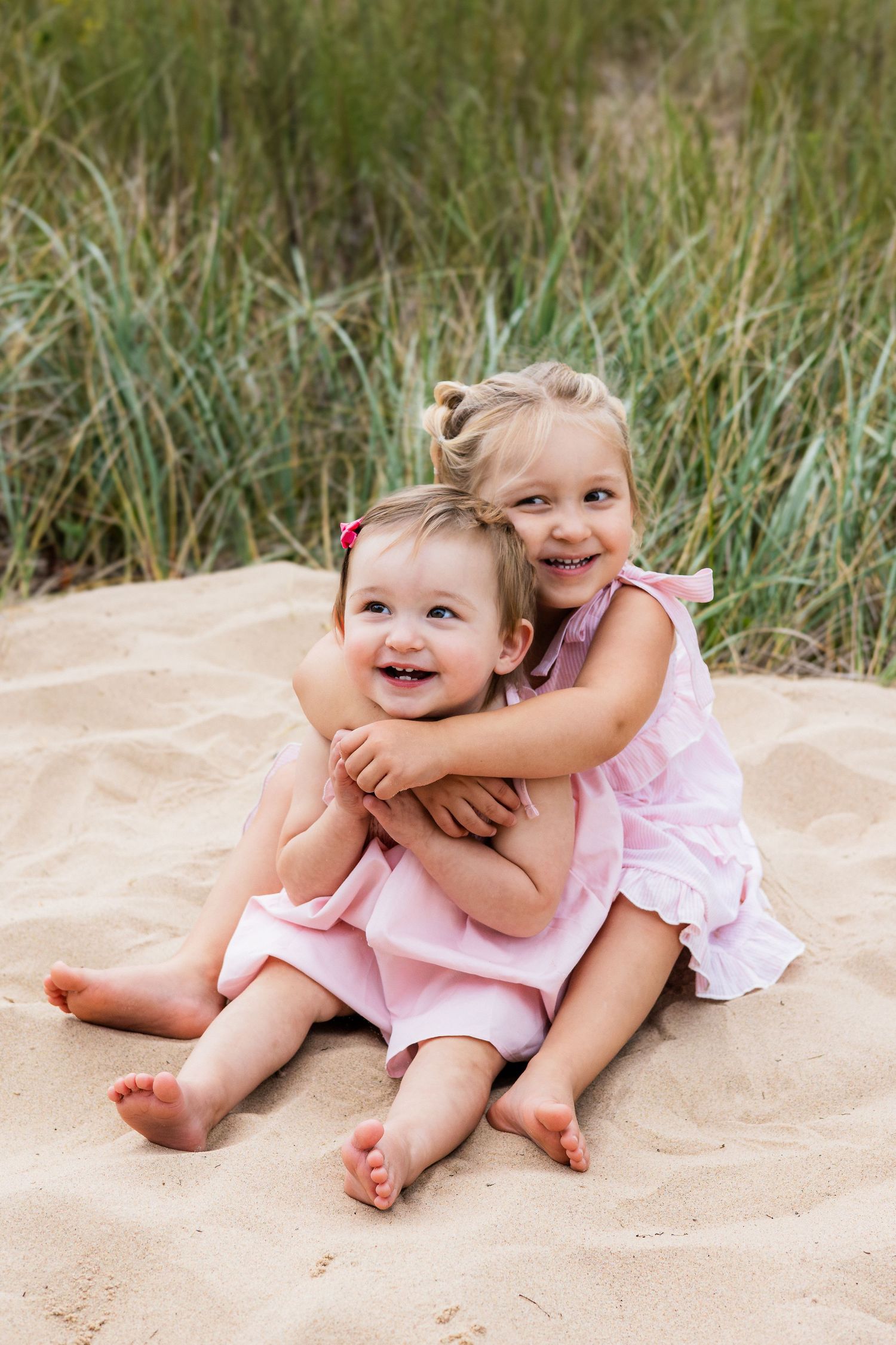 Two young sisters in pink dresses sit together on sandy beach, older girl hugging younger toddler, both smiling happily.