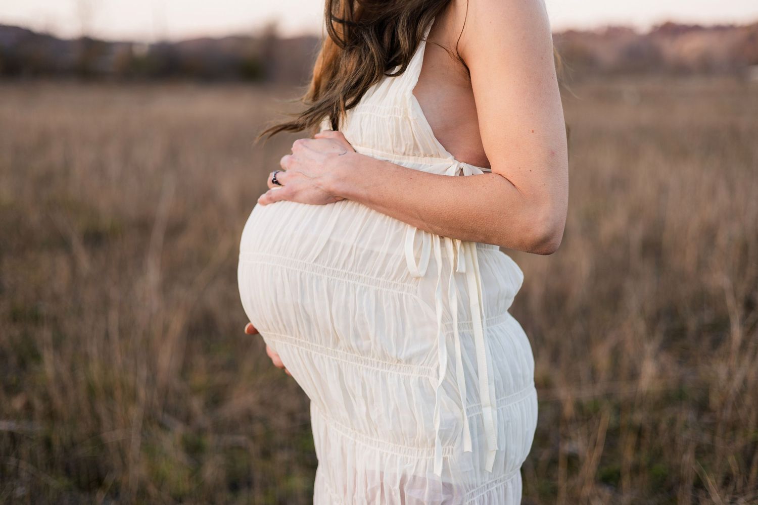 Pregnant woman in white dress cradling baby bump in golden field during maternity photo session.