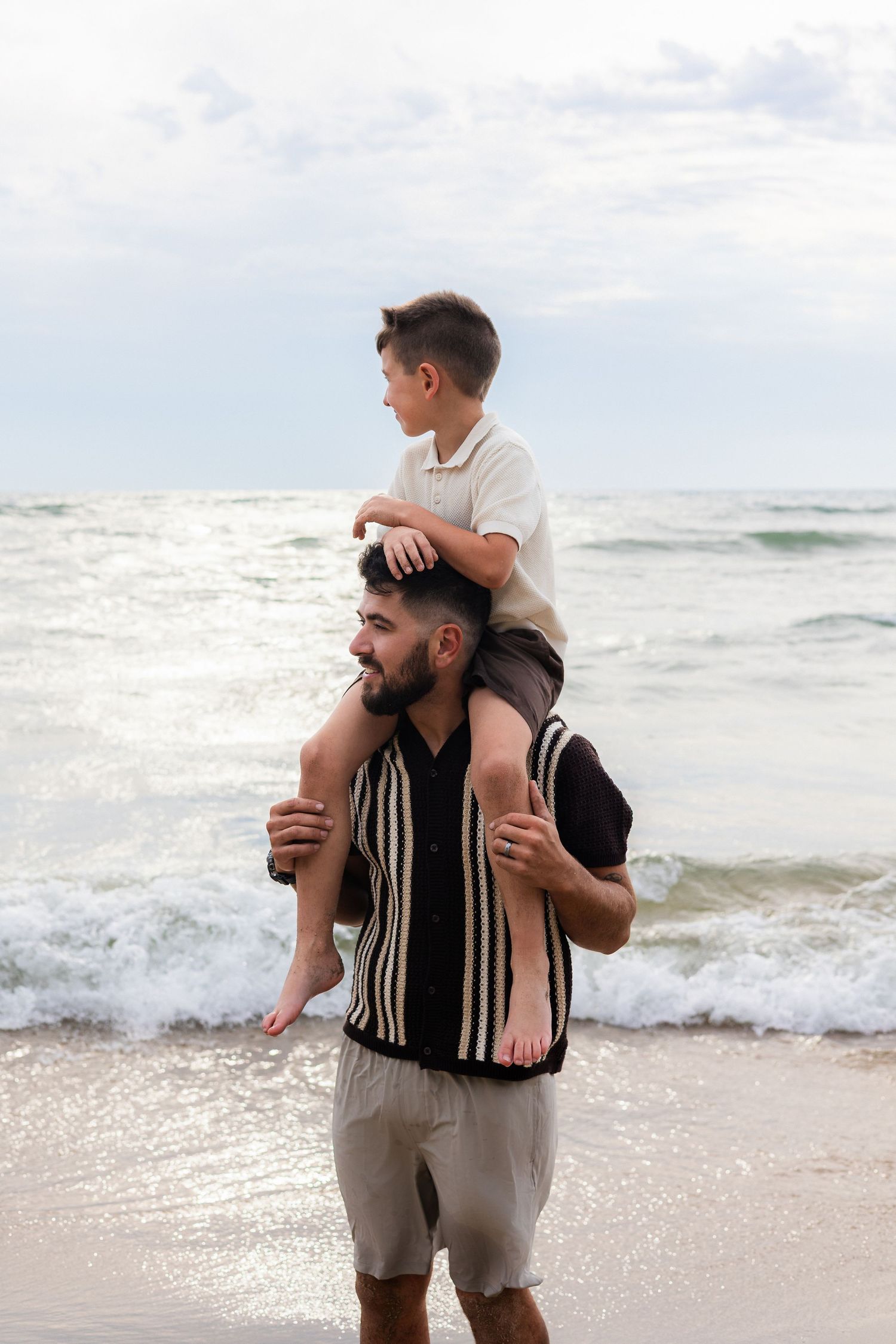 Father carries young son on his shoulders while standing on a sandy beach with ocean waves behind them.