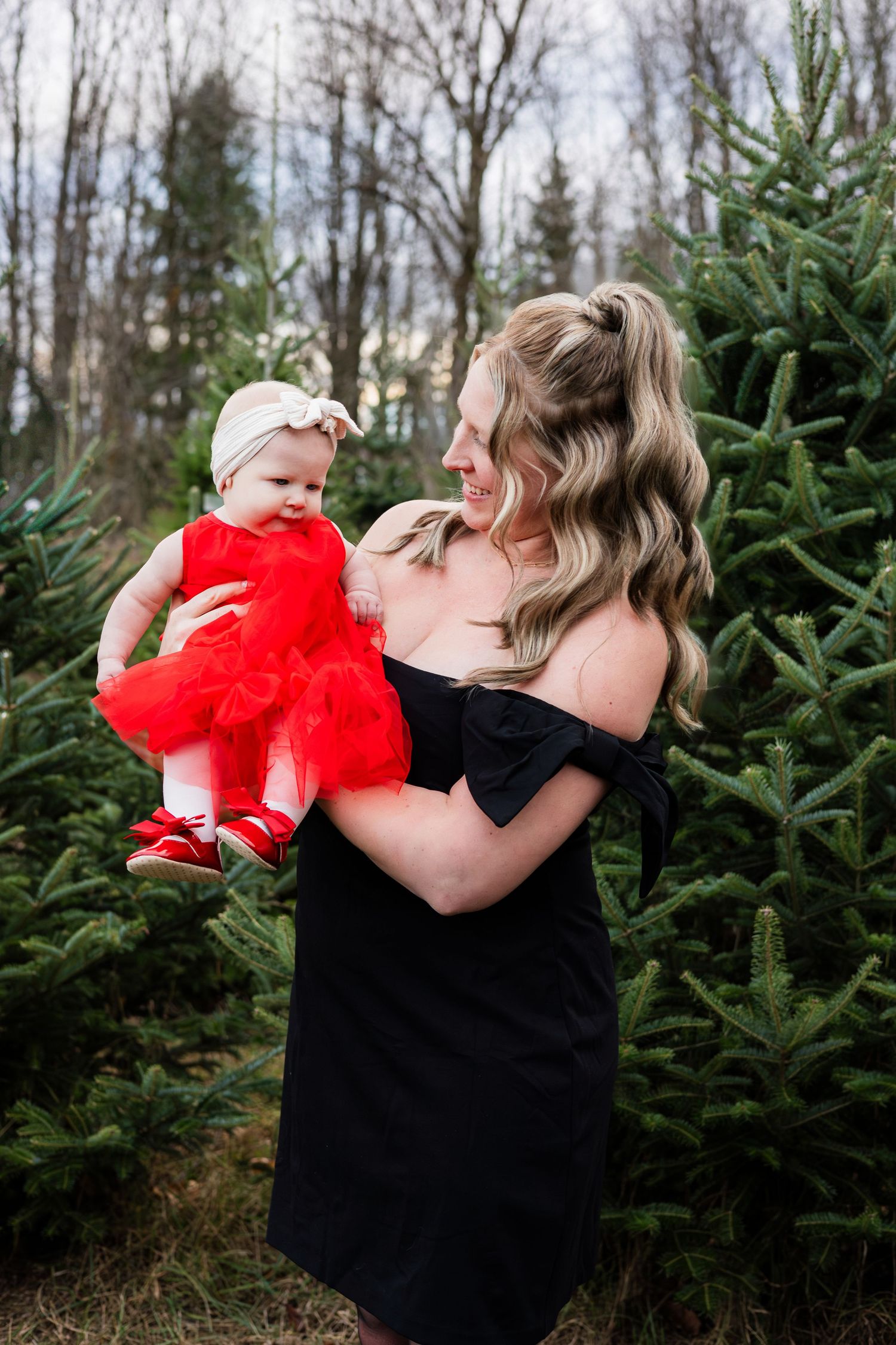 Mother in black off-shoulder dress holding baby girl in red tulle dress at a Christmas tree farm.