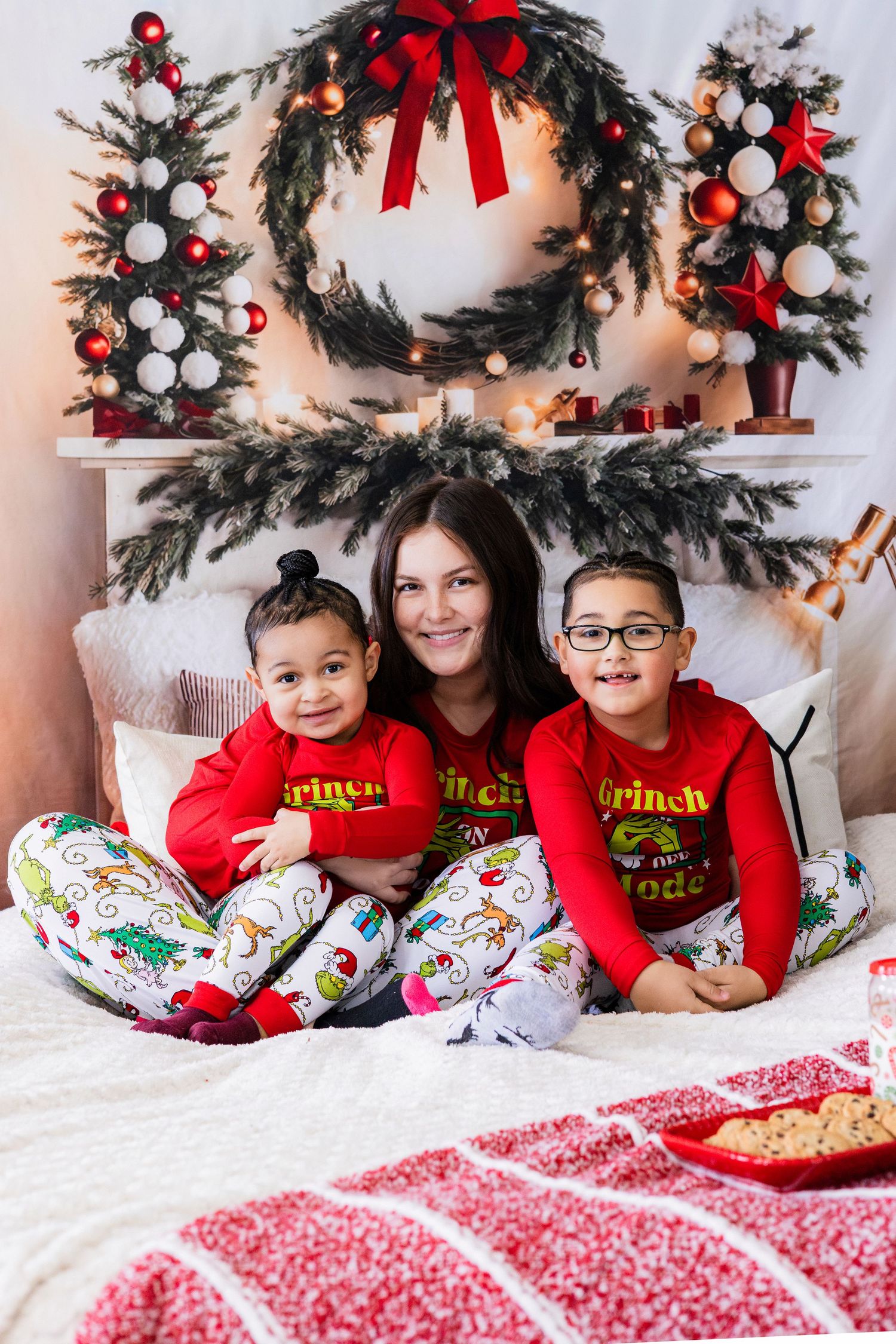 Family of three in matching Grinch Christmas pajamas sitting on bed with festive holiday decorations.
