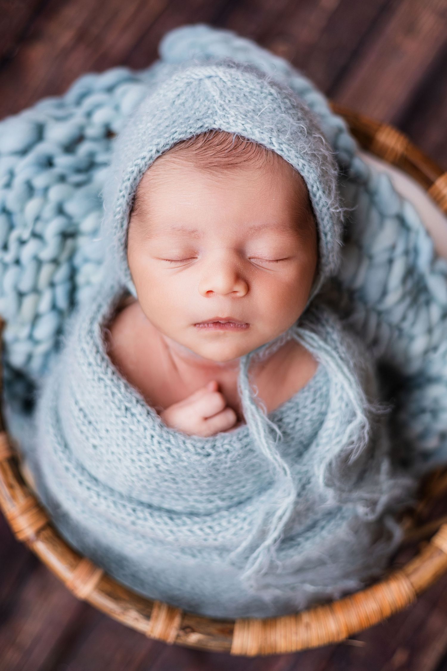 Newborn baby sleeping in a wicker basket, swaddled in a soft blue knit wrap and matching bonnet.