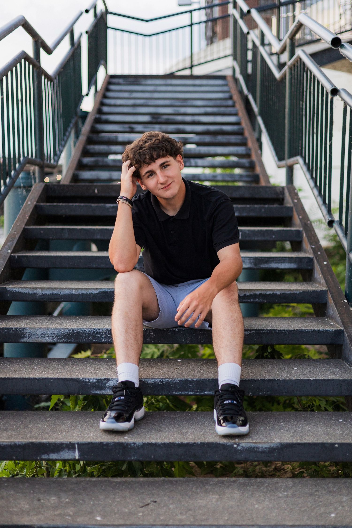 Teen boy in black t-shirt and gray shorts sitting casually on outdoor metal stairs with hand resting on his head.