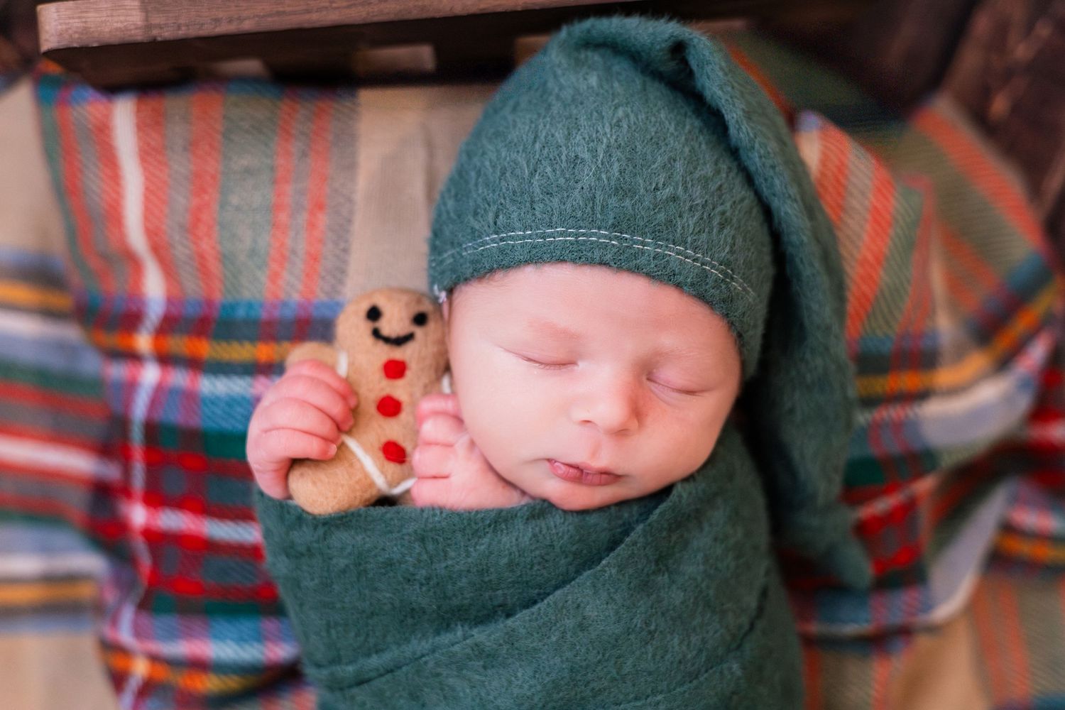 Sleeping newborn baby in teal knit wrap and hat, holding a small gingerbread man toy ornament.