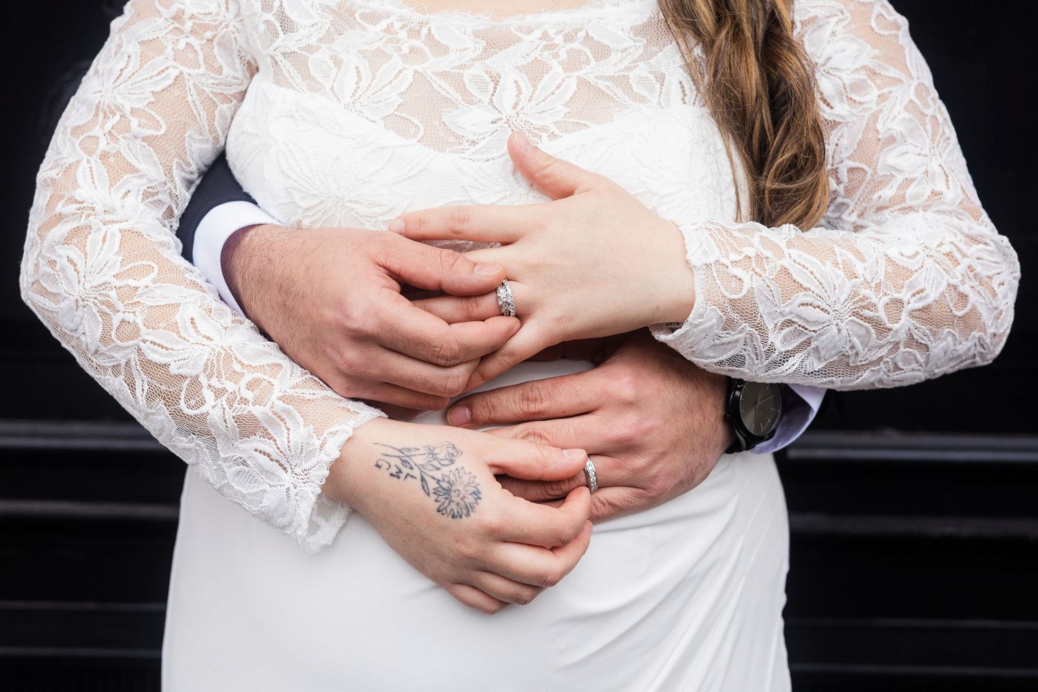 Bride and groom holding hands showing wedding rings, bride wearing lace long-sleeve wedding dress with floral tattoo visible.