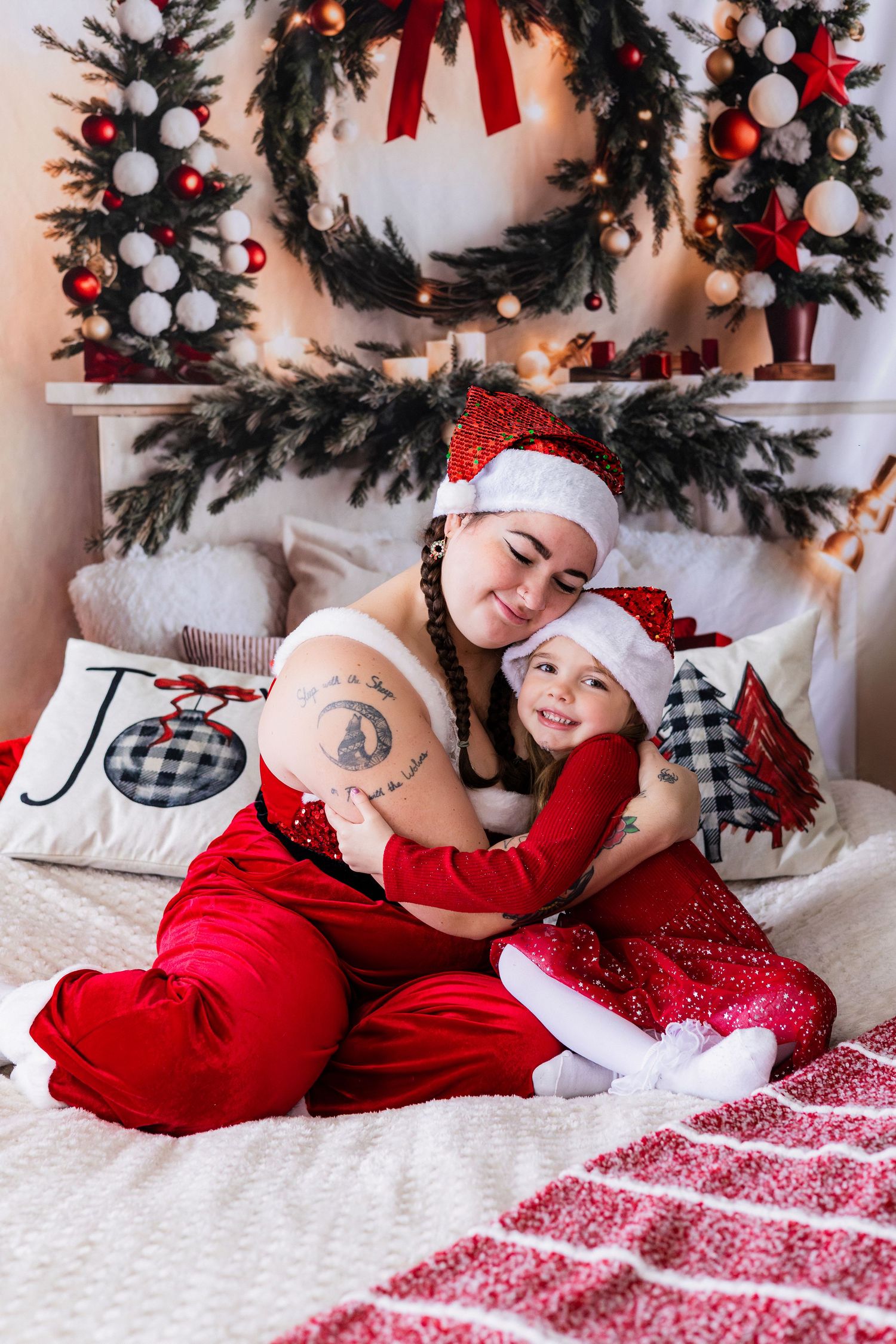 Mother and daughter in matching red Santa outfits hugging on bed with Christmas decor.