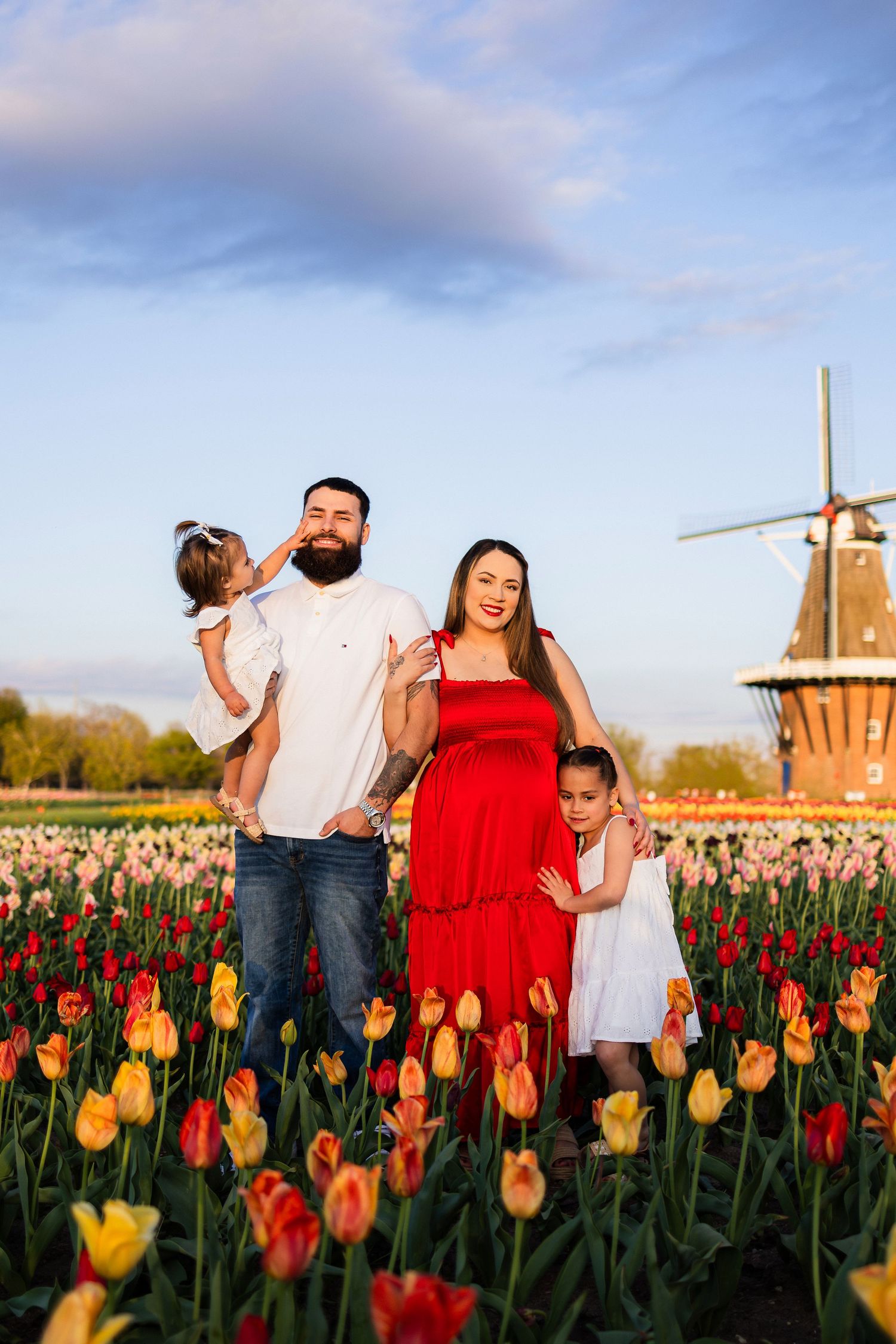 Family posing in vibrant tulip field with windmill backdrop at sunset, woman in red dress, man holding toddler, girl in white.