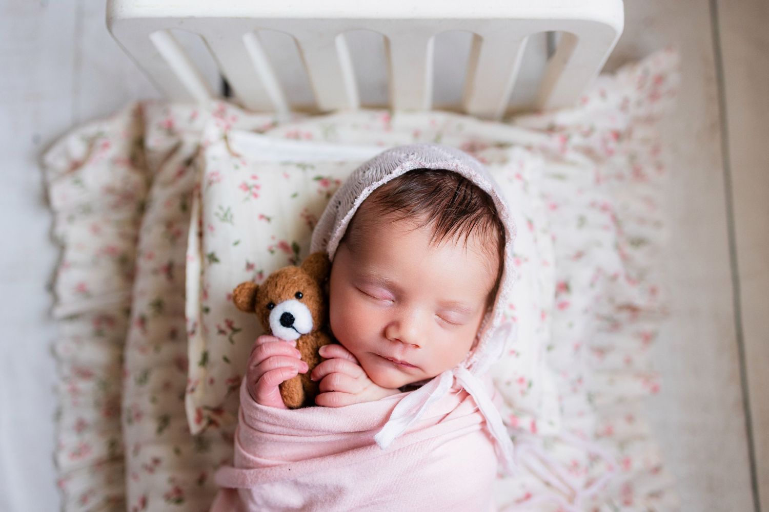 Sleeping newborn baby girl swaddled in pink holding a small teddy bear in a white crib.