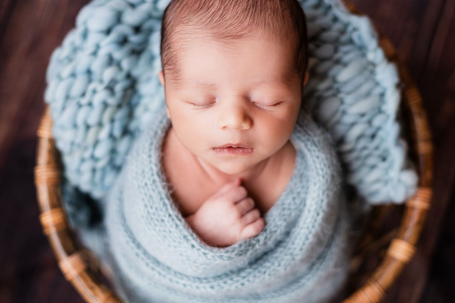 Sleeping newborn baby wrapped in light blue knit blanket, resting peacefully in a wicker basket.