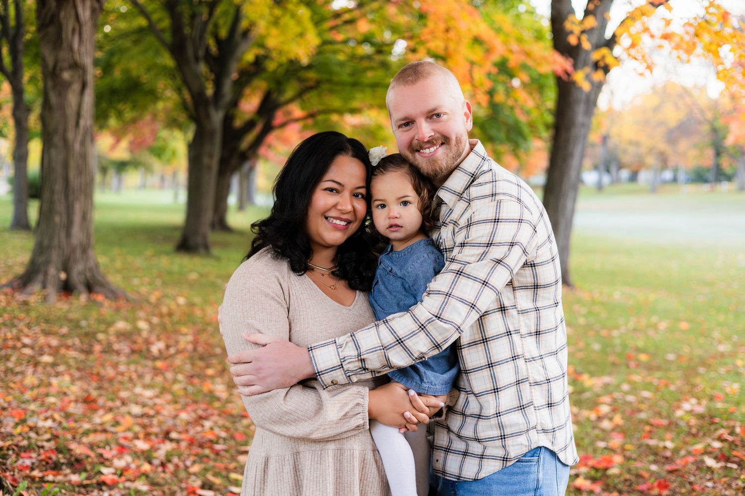 Family of three posing outdoors among colorful autumn foliage, smiling couple holding young toddler in a park setting.