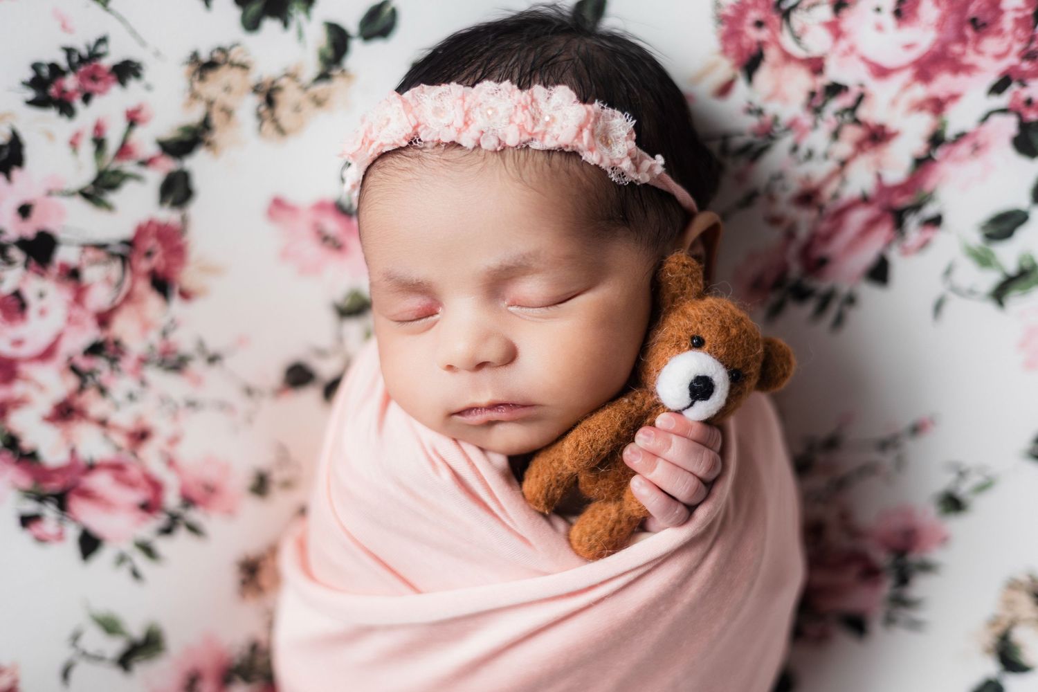 Sleeping newborn baby girl swaddled in pink wrap holding tiny teddy bear on floral background with pink headband.