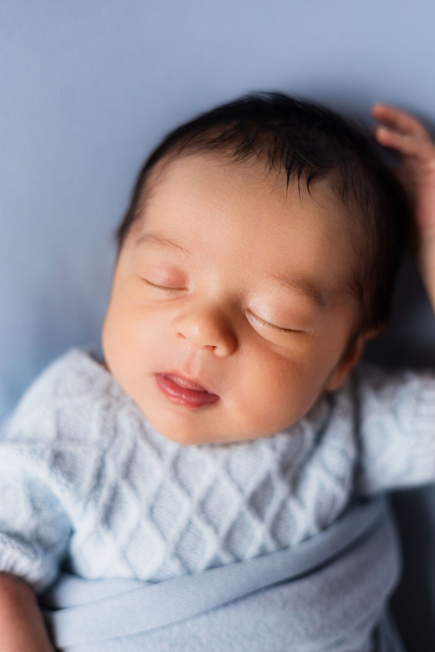 Peaceful sleeping newborn baby boy in blue cable-knit outfit, wrapped in blue swaddle on blue background.