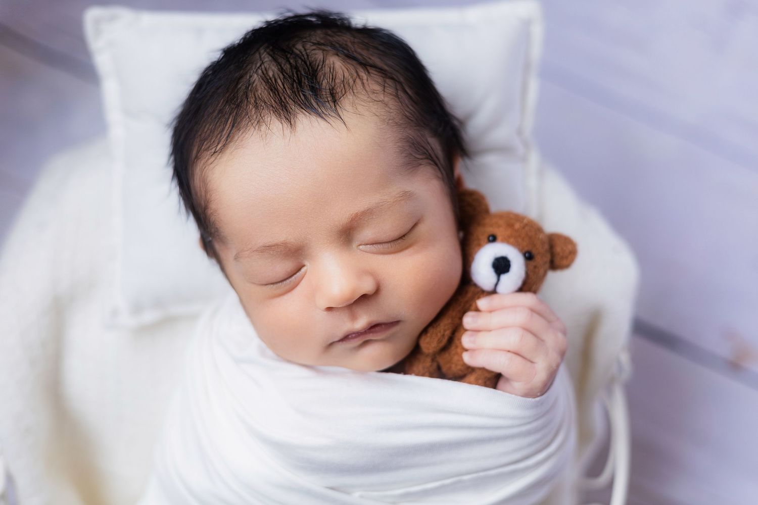 Peaceful newborn baby sleeping swaddled in white wrap, holding a tiny brown teddy bear on a white pillow.