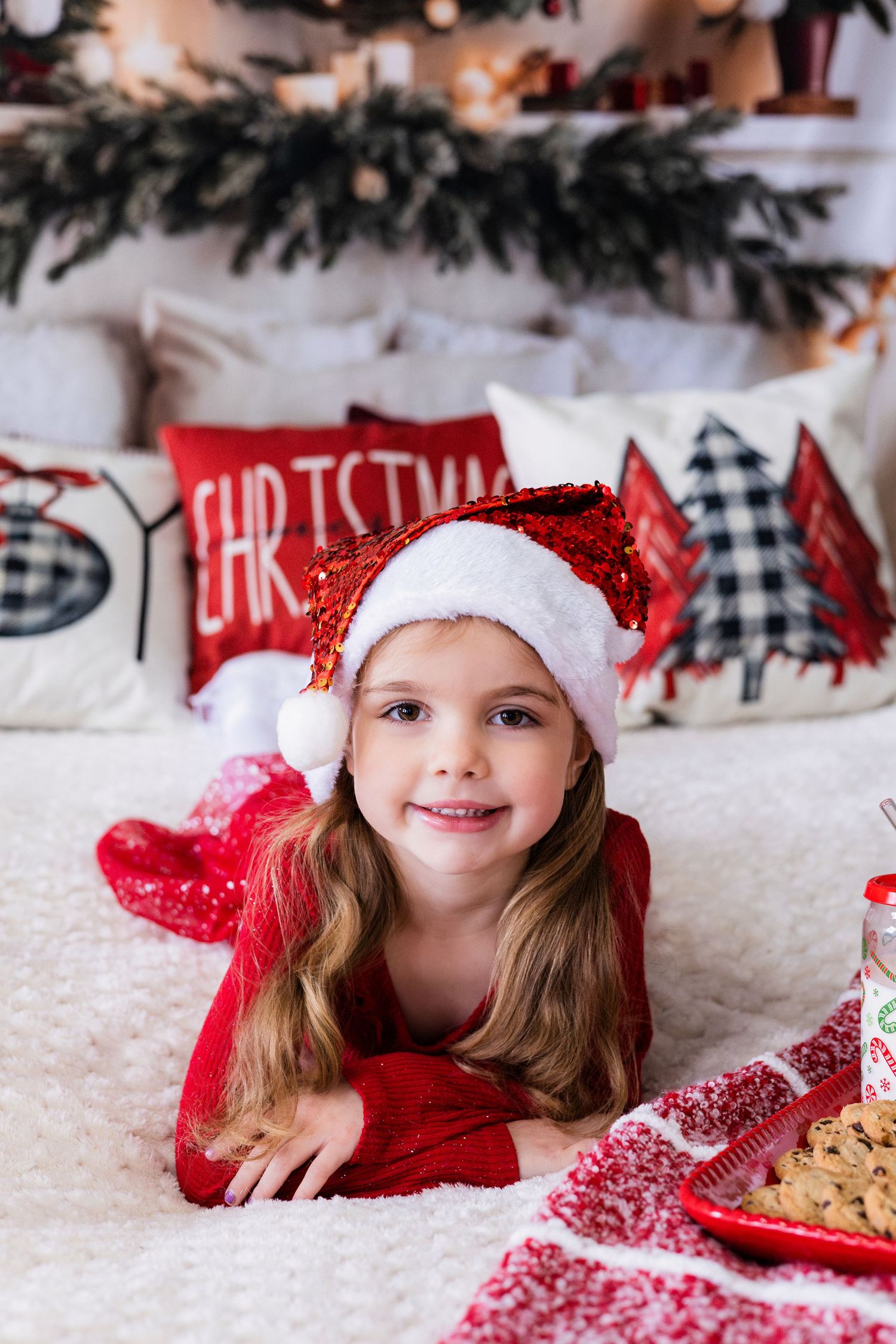 Cute little girl in red dress and Santa hat lying on bed with Christmas decorations.