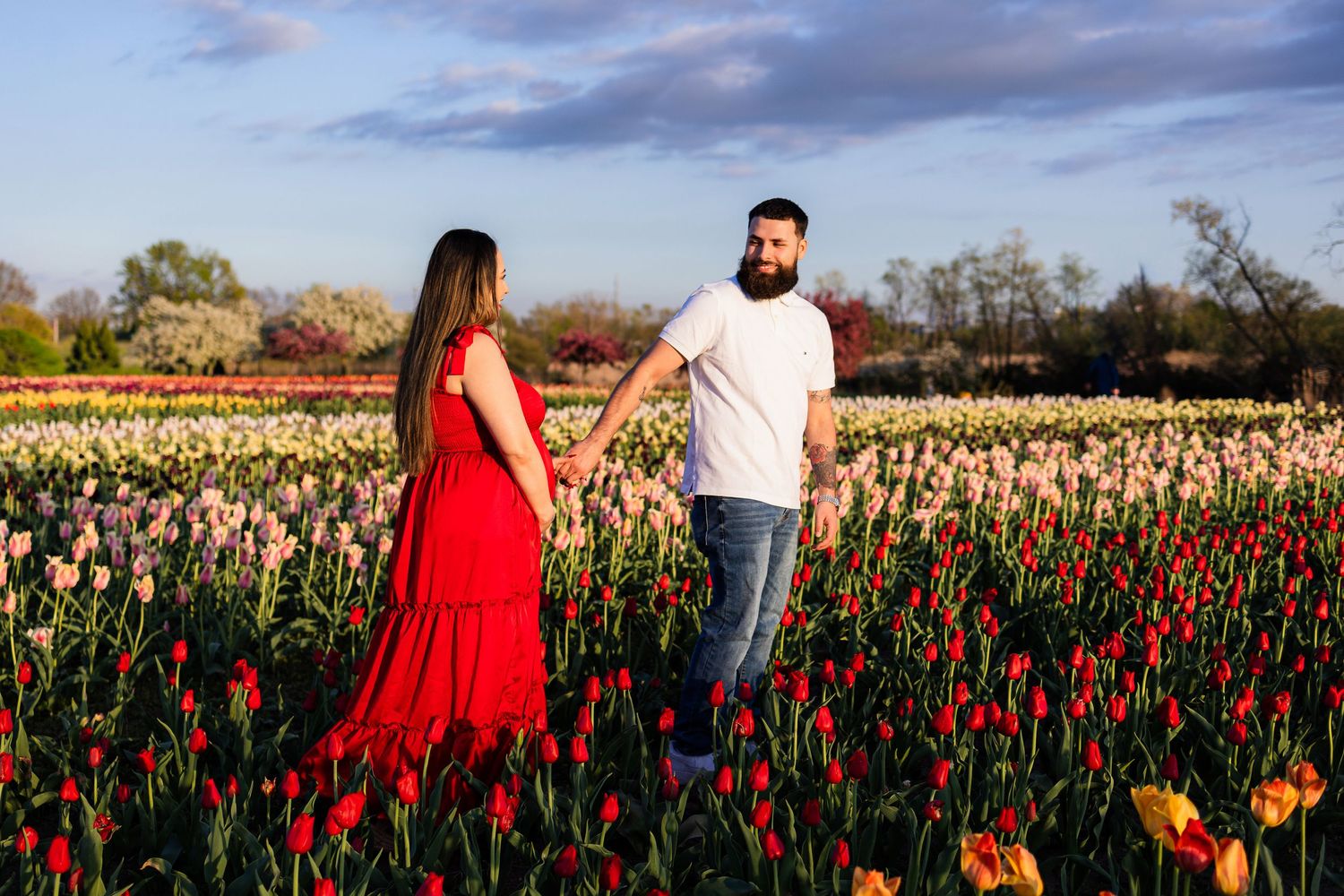 Couple holding hands in a vibrant tulip field, woman in red dress, man in white shirt, colorful blooms under blue sky.