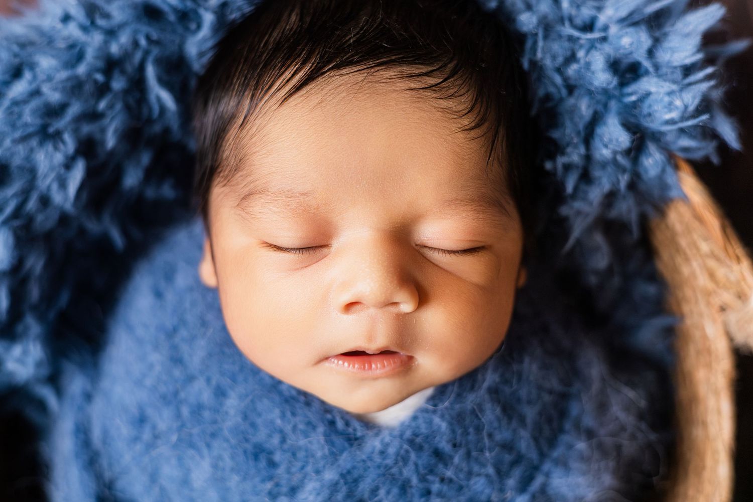 Peaceful newborn baby sleeping wrapped in a blue fuzzy blanket in a basket.