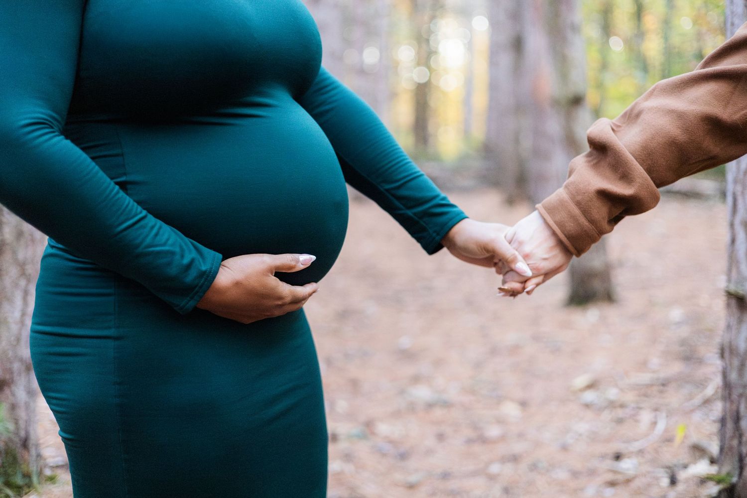 Pregnant woman in teal dress holding belly while partner holds her hand in an autumn forest setting.