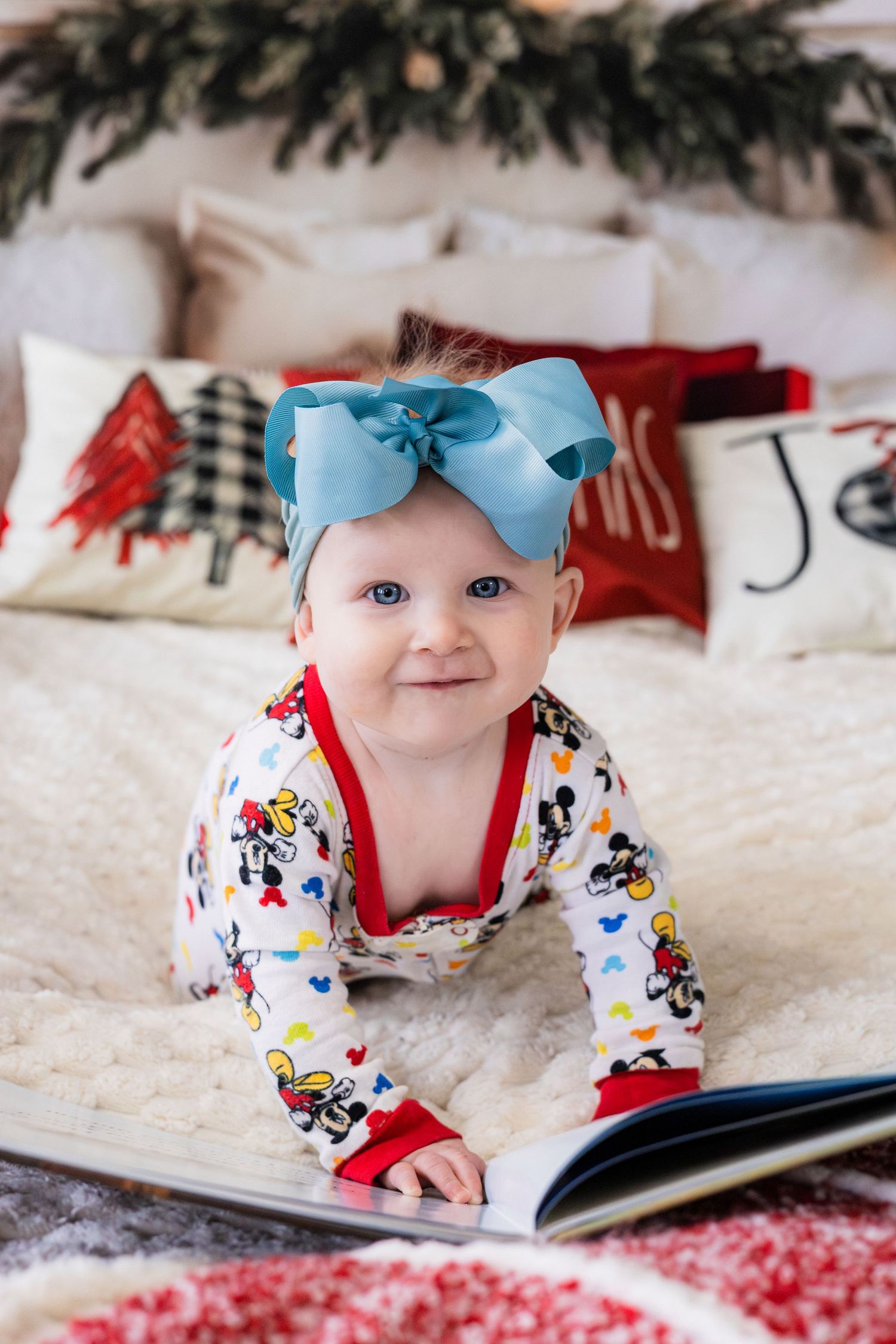 Adorable baby girl with blue bow headband in Mickey Mouse pajamas reading a book on Christmas bed.