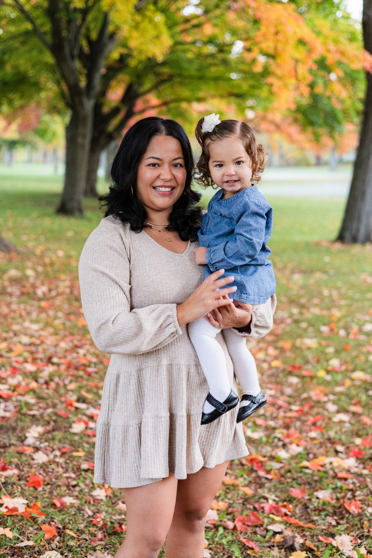 Smiling mother holding her toddler daughter in a denim outfit among colorful autumn leaves in a park.