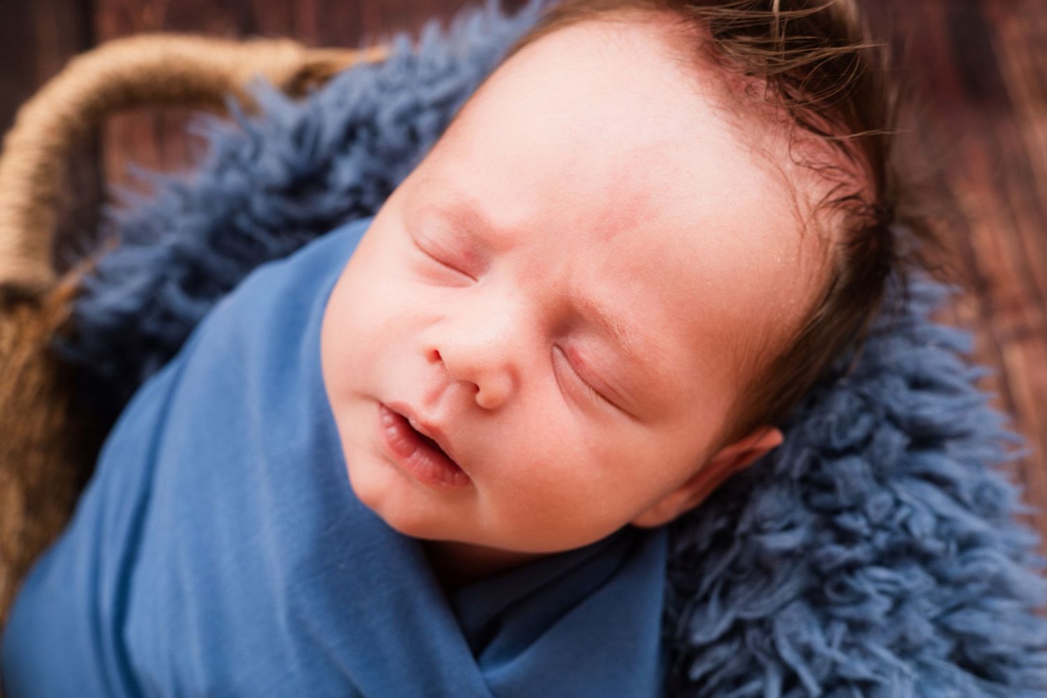 Sleeping newborn baby swaddled in a blue wrap, resting on a soft blue fuzzy blanket in a basket.