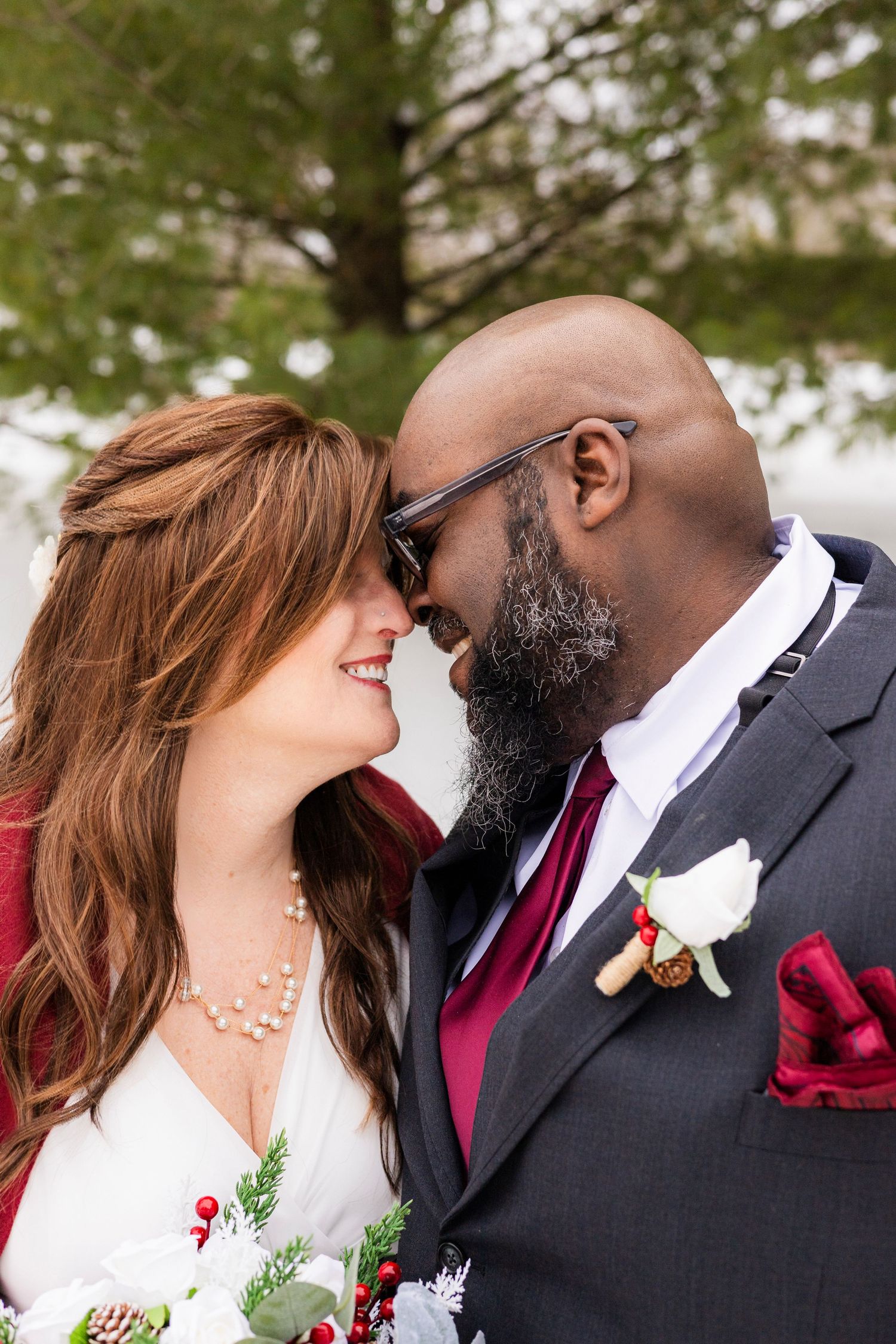 Interracial newlywed couple sharing romantic moment, groom in gray suit with burgundy tie and boutonniere, bride holding bouquet.