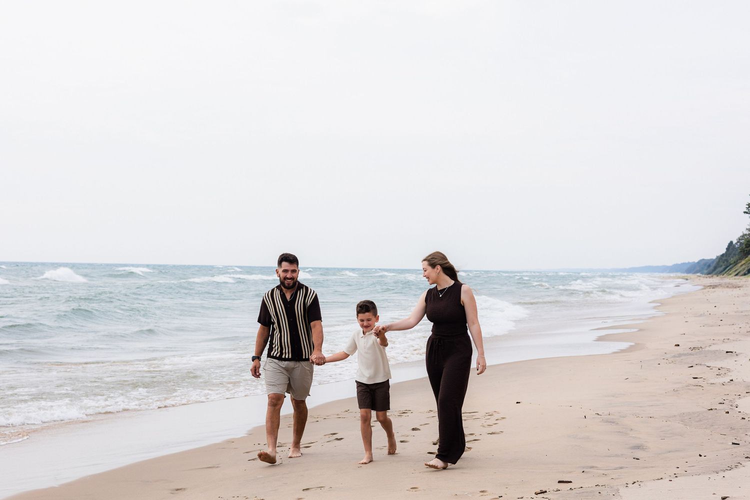 Family of three walking hand-in-hand on a sandy beach near ocean waves on a cloudy day.