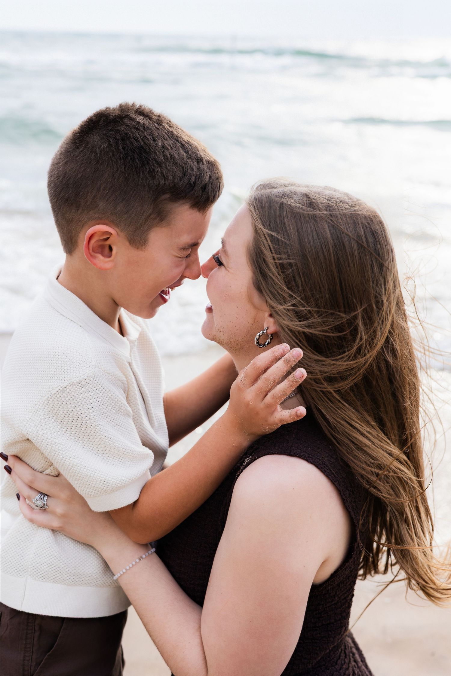 Mother and son share a joyful nose-to-nose moment on a beach with ocean waves in the background.