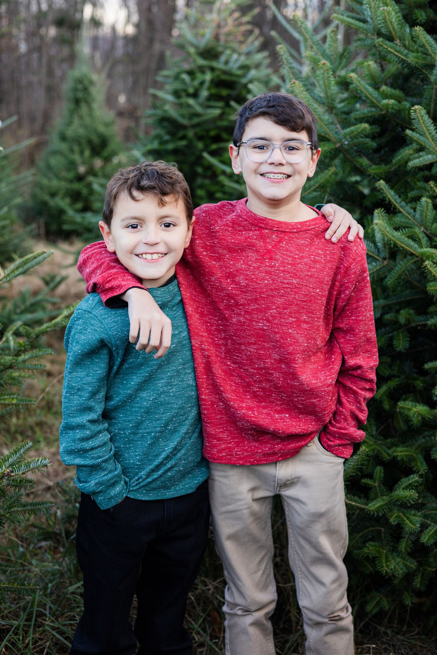 Two young brothers smiling together at a Christmas tree farm, wearing red and teal sweaters in a holiday portrait.