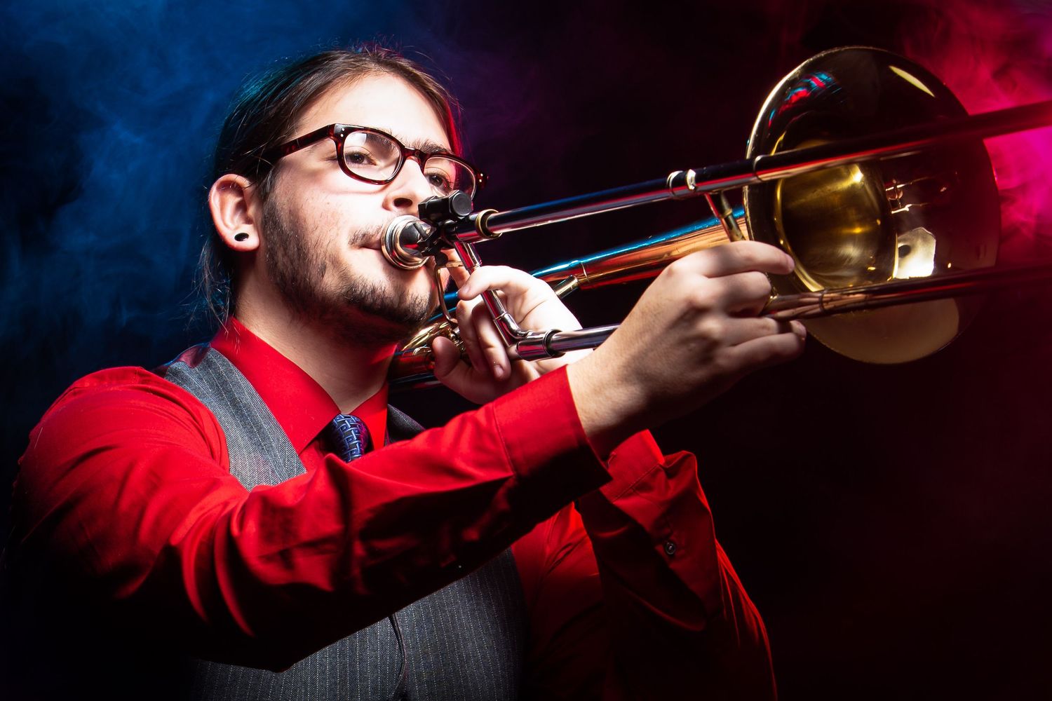 A musician in a red shirt plays trombone on stage under dramatic red and blue lighting during a performance.