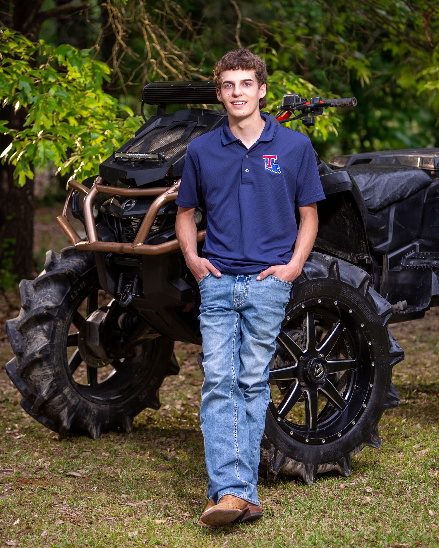 A person in a navy blue polo shirt and jeans poses beside a large black tractor tire in an outdoor setting.