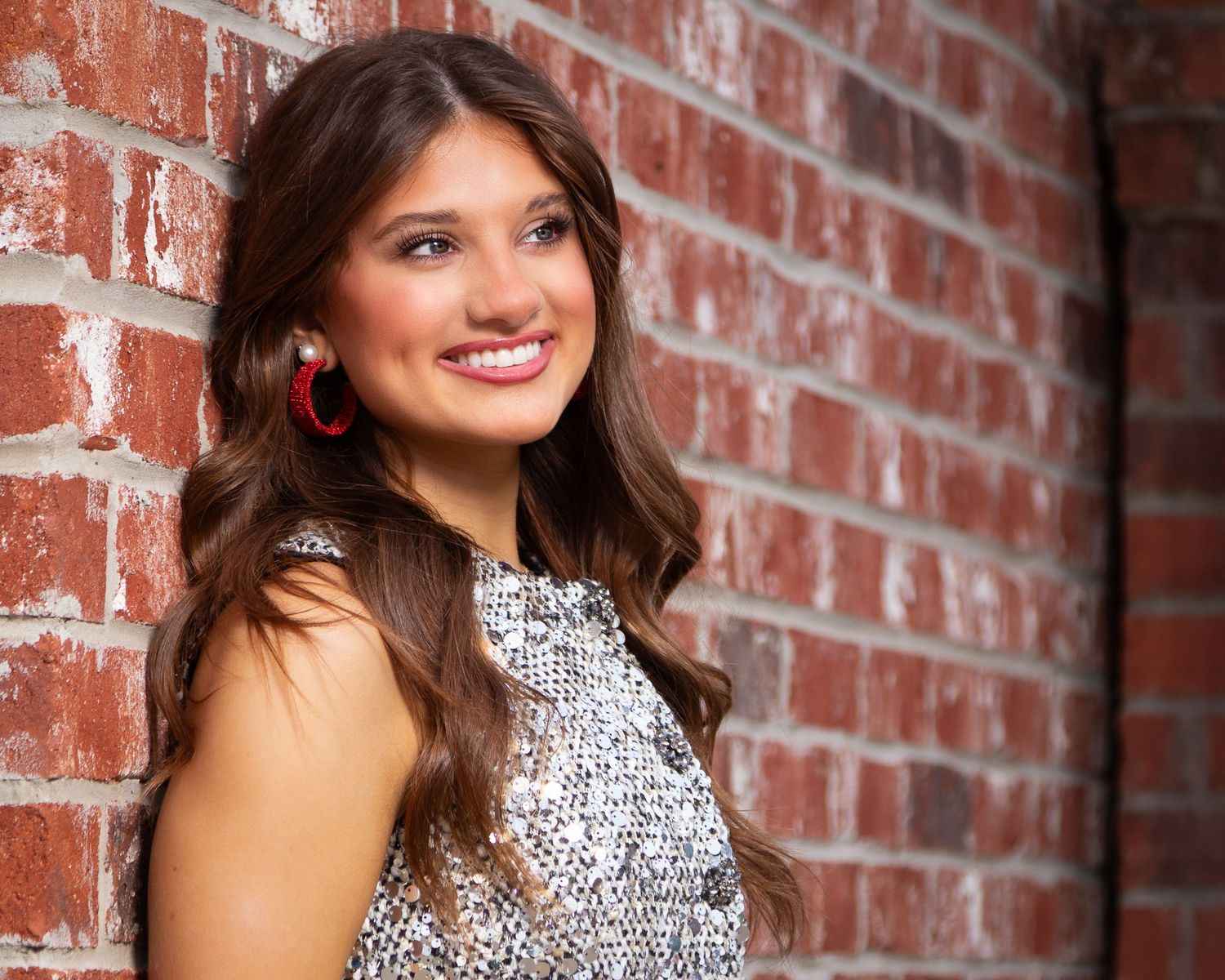 A person in a sequined dress smiles while posing against a rustic brick wall background.
