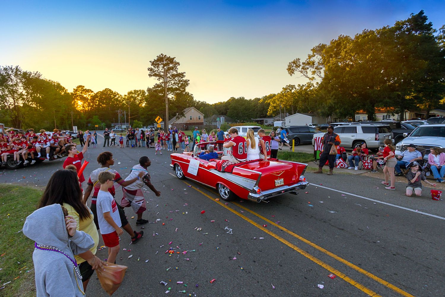 A classic red convertible car leads a festive parade at dusk, with crowds lining the street under tall trees.