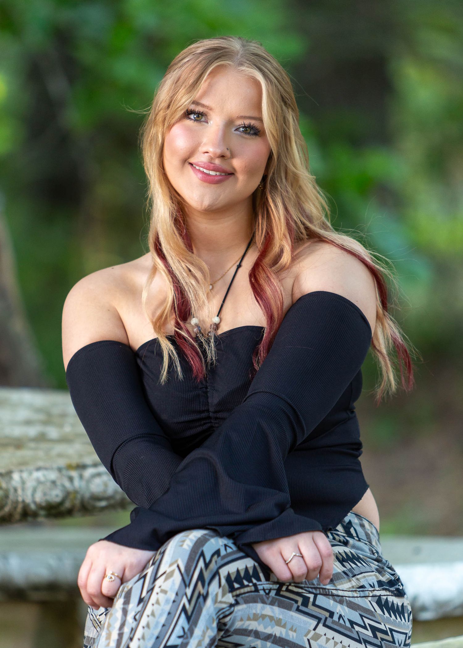 A high school senior in a black sweater and patterned skirt poses outdoors against a blurred natural background.