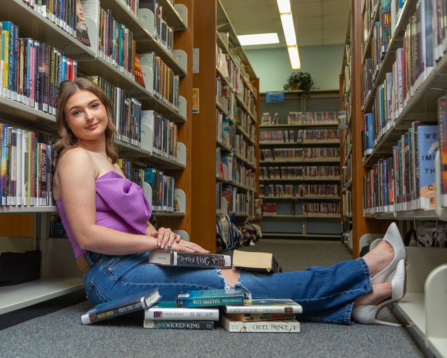 Student in purple top reclining between library bookshelves with textbooks and study materials.