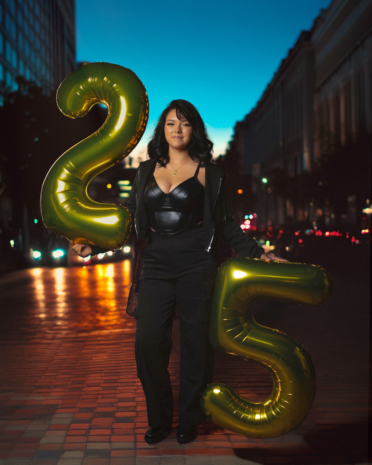 A person in black clothing poses with large metallic gold number balloons against an urban evening backdrop.