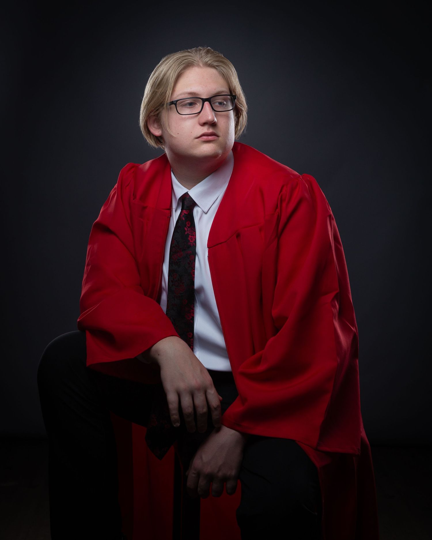 A figure in a red academic gown and white shirt sits against a dark background in a dramatic portrait series.