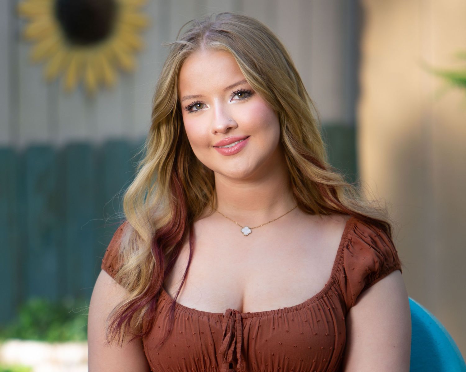 A high school senior in a brown top smiles warmly while posing for a professional outdoor portrait session.