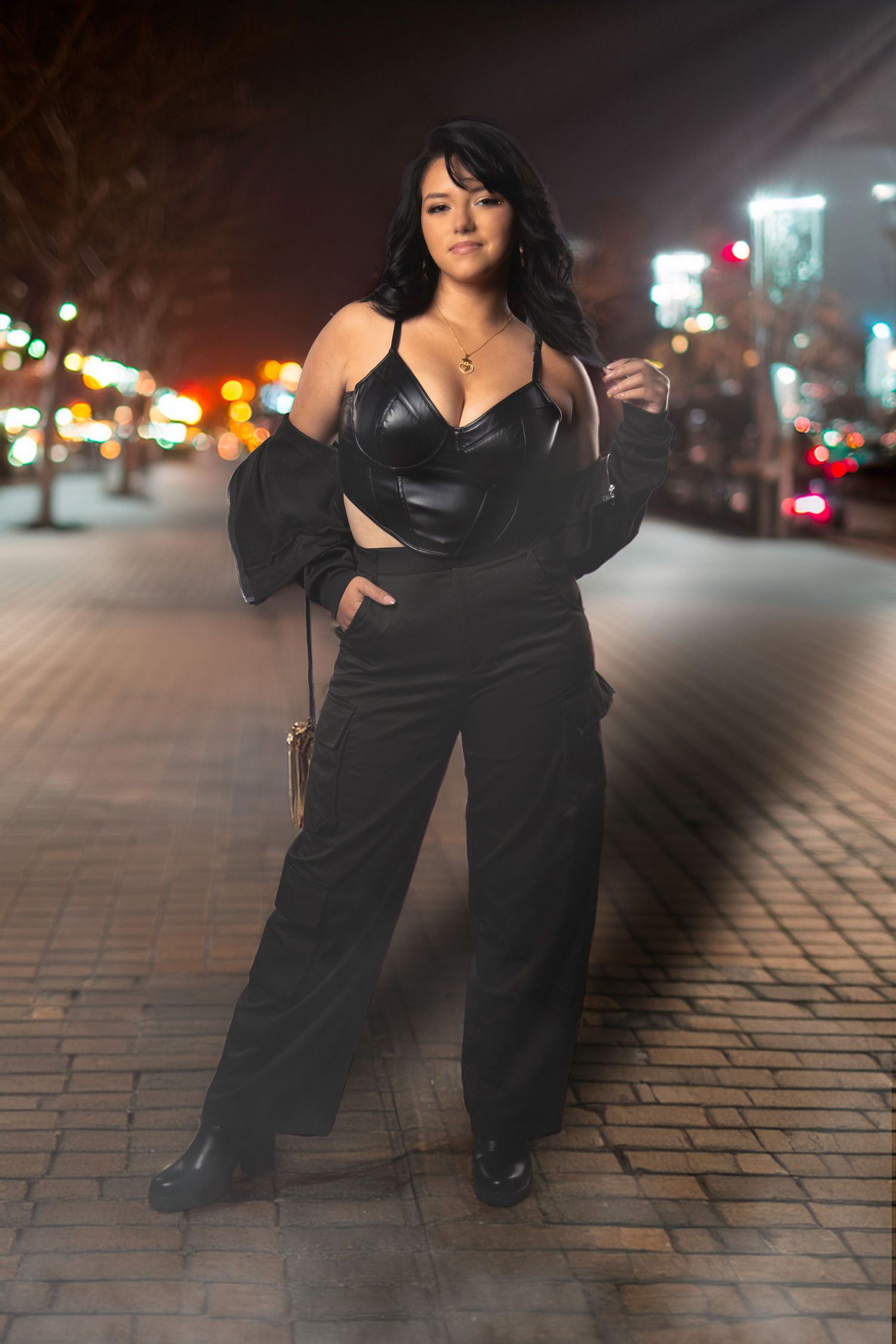 A figure in a flowing black dress poses dramatically on brick pavement at night with city lights in the background.